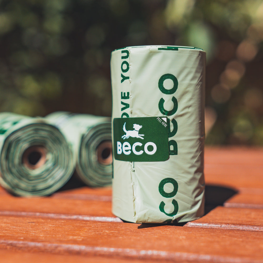 Rolls of Beco brand compostable poo bags placed on a wooden surface with greenery in the background.