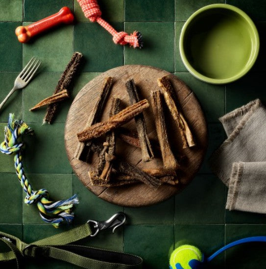 Dog treats on a wooden plate with toys and a bowl on a green tiled floor.